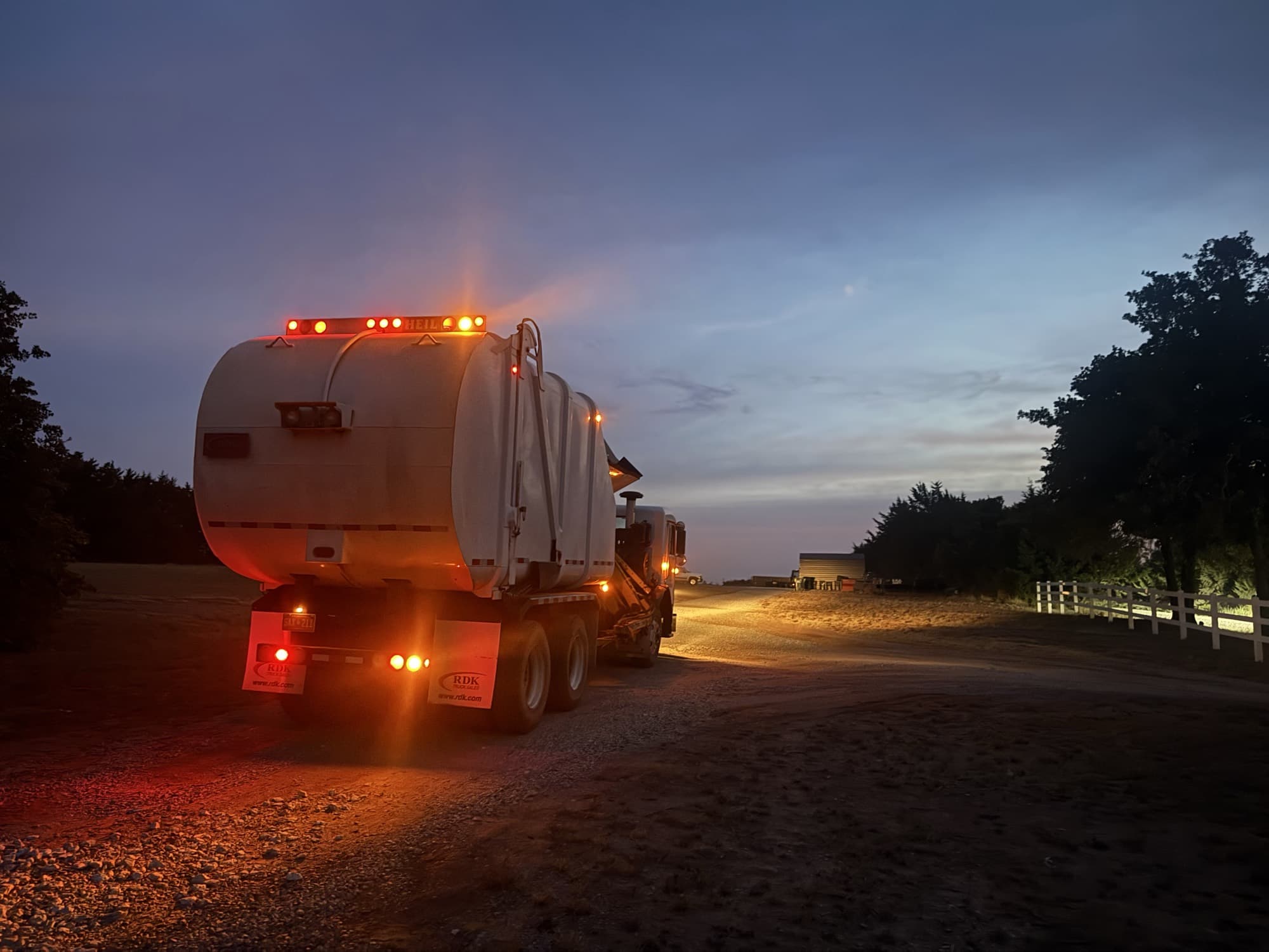A DHH Disposal truck heading out on a gravel road at dawn, running lights glowing